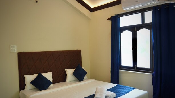 A hotel room with a bed, a window with curtains, an air conditioning unit, and a wall-mounted security camera.