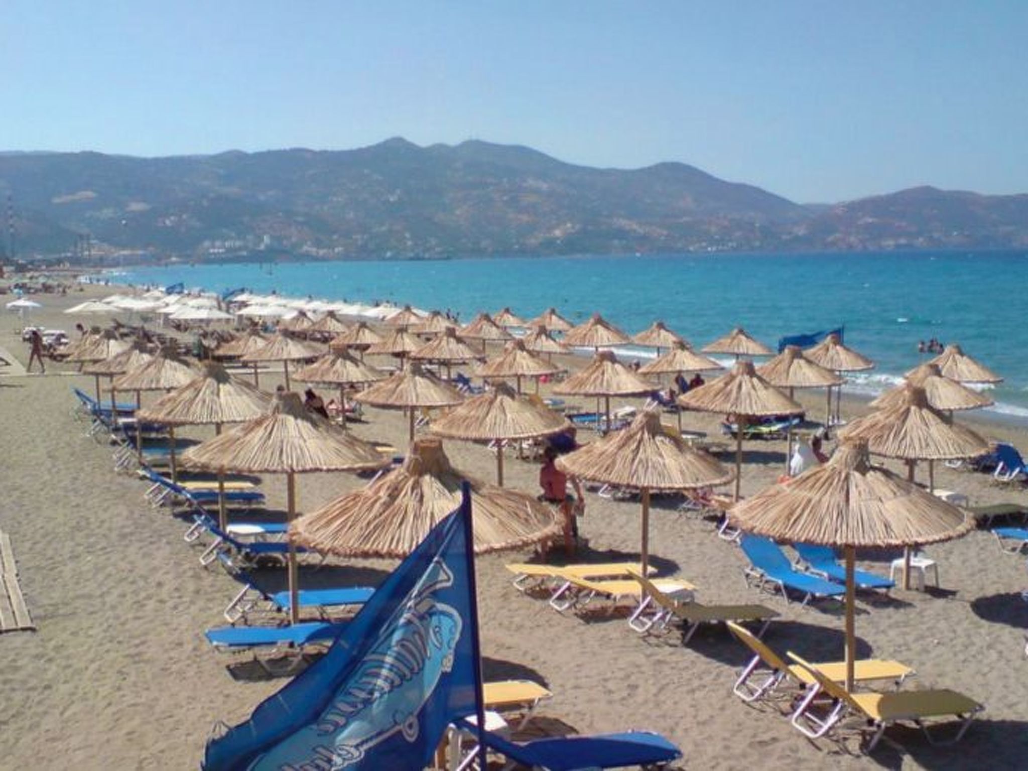A beach with rows of sun umbrellas and lounge chairs, a clear sky, and a mountainous backdrop.
