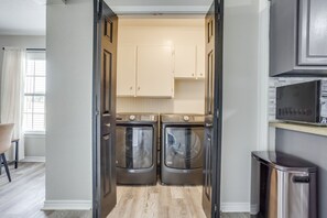 A laundry room with two front-loading washing machines, a microwave above, and a trash can to the right.