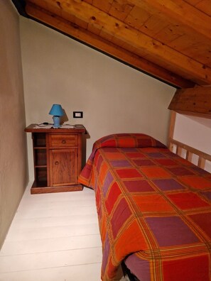 A bedroom with a wooden headboard, a plaid bedspread, a nightstand with a lamp, and a wall socket. - Relaxing Home with a View of the Sea (Toscana)