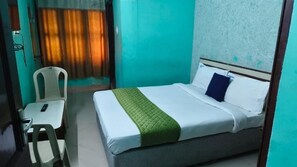 Habitación individual con una colcha verde, una mesita de noche con un control remoto y una silla.