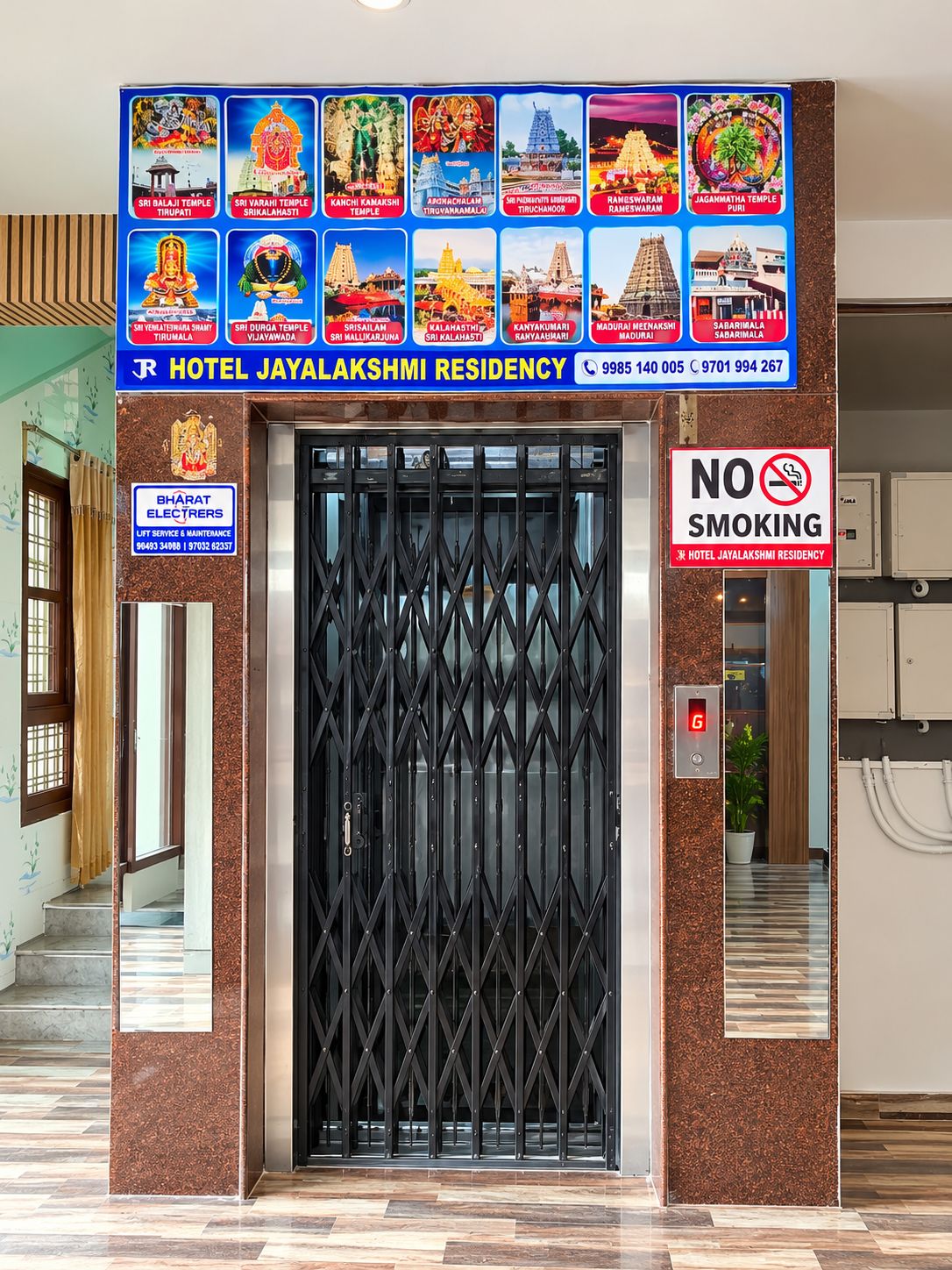 La entrada del Hotel Jayalakshmi Residency, con un cartel de prohibido fumar, un ascensor y una señalización que muestra varios templos.