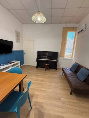 Un salón con una mesa de madera, sillas azules, un piano negro, un sofá marrón con cojines azules, un televisor en la pared y una lámpara colgante.