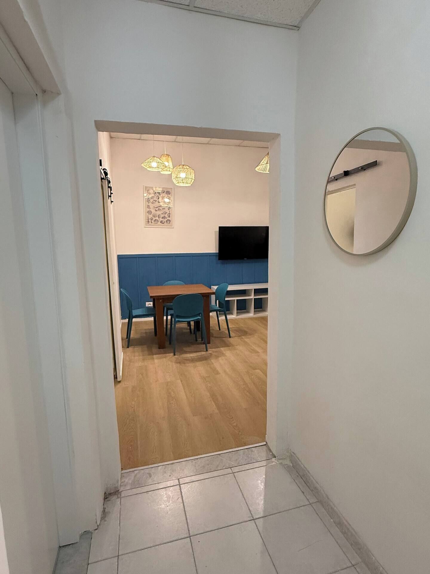 Un couloir mène à une salle à manger avec un mur bleu, une table en bois et des chaises. Un téléviseur à écran plat est fixé au mur, et un miroir rond se trouve sur le côté droit.