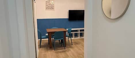 Un couloir mène à une salle à manger avec un mur bleu, une table en bois et des chaises. Un téléviseur à écran plat est fixé au mur, et un miroir rond se trouve sur le côté droit.