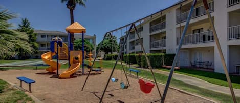 Un terrain de jeux avec des balançoires et un toboggan, entouré d’un bâtiment doté de balcons.