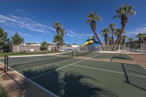 An outdoor tennis court with a net, surrounded by palm trees and a water slide structure in the background. - Las Palmas 1605 | Next to Clubhouse and Pool! (Saint George)