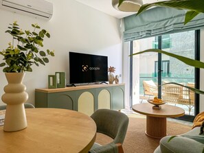 Ein modernes Wohnzimmer mit Flachbildfernseher, Esstisch, bequemen Sitzmöglichkeiten, einer Topfpflanze und Blick ins Freie.