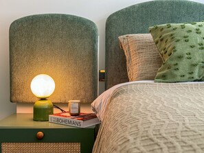 Ein ordentlich bezogenes Bett mit grünem Kopfteil, einer Nachttischlampe und einem Buch mit dem Titel 'The New Bohemians' auf dem Nachttisch.