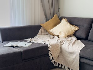 A grey sofa with a beige throw blanket and two pillows, one with a woven texture and the other plain. An open book is on the sofa. - Qoople Legend 37 А-30 100 Meter vom Strand entfernt by Interhome (Alanya)