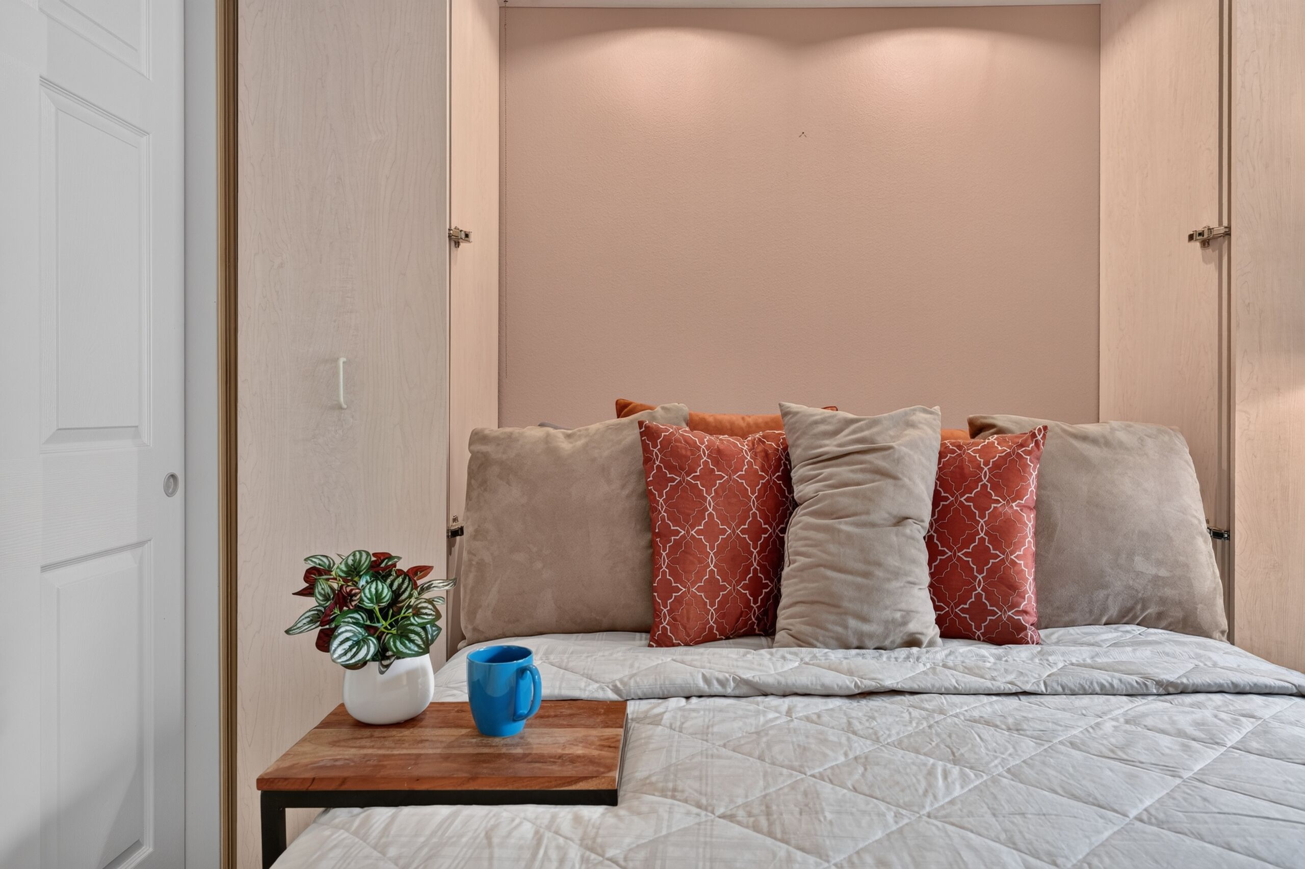 Un dormitorio con una cama, almohadas, una mesita de noche con una planta y una taza, y una puerta.