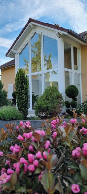 Une maison dotée d’un grand porche vitré, entourée d’arbustes bien entretenus et de plantes à fleurs.