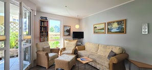 Ein Wohnzimmer mit einer Couch, zwei Sesseln, einem Couchtisch, einem Bücherregal, einem Fernseher und einem Fenster mit Blick auf Grünflächen.