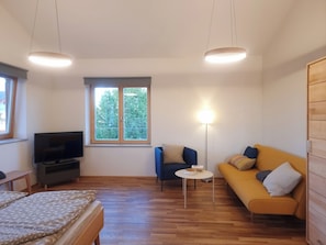 A modern living room with a flat-screen TV, a sofa, a small round table, and a window with a view of trees. - Vacation Rental Next to the Ceramics Studio (Babensham)