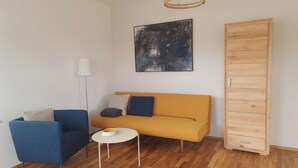 Een moderne woonkamer met een bank, stoelen, een bijzettafeltje, een vloerlamp, een schilderij en een houten garderobe.
