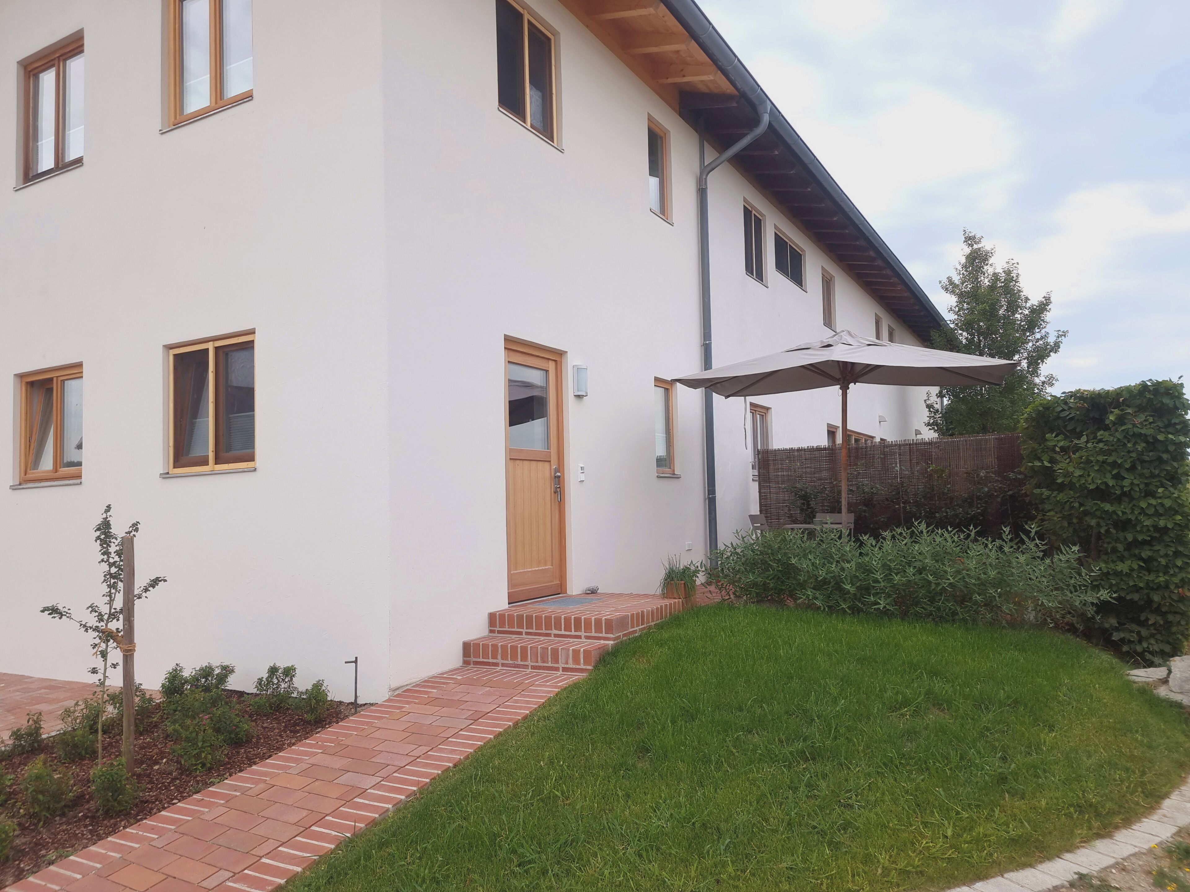 Una moderna casa a due piani con un vialetto in mattoni che conduce all'ingresso, una porta in legno e un piccolo giardino con una panchina e un ombrellone.