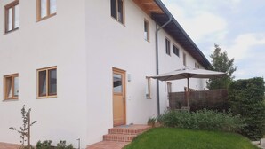 Een modern huis met twee verdiepingen, een pad van baksteen naar de ingang, een houten deur en een kleine tuin met een bankje en parasol.