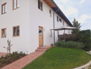 A modern two-story house with a brick pathway leading to the entrance, a wooden door, and a small garden area with a bench and umbrella. - Vacation Rental Next to the Ceramics Studio (Babensham)