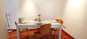 Ein Essbereich mit einem weißen Tisch, gedeckt für zwei Personen, zwei orangefarbenen Stühlen, einer Vase mit einer roten Blume und einer Heizung an der Wand.
