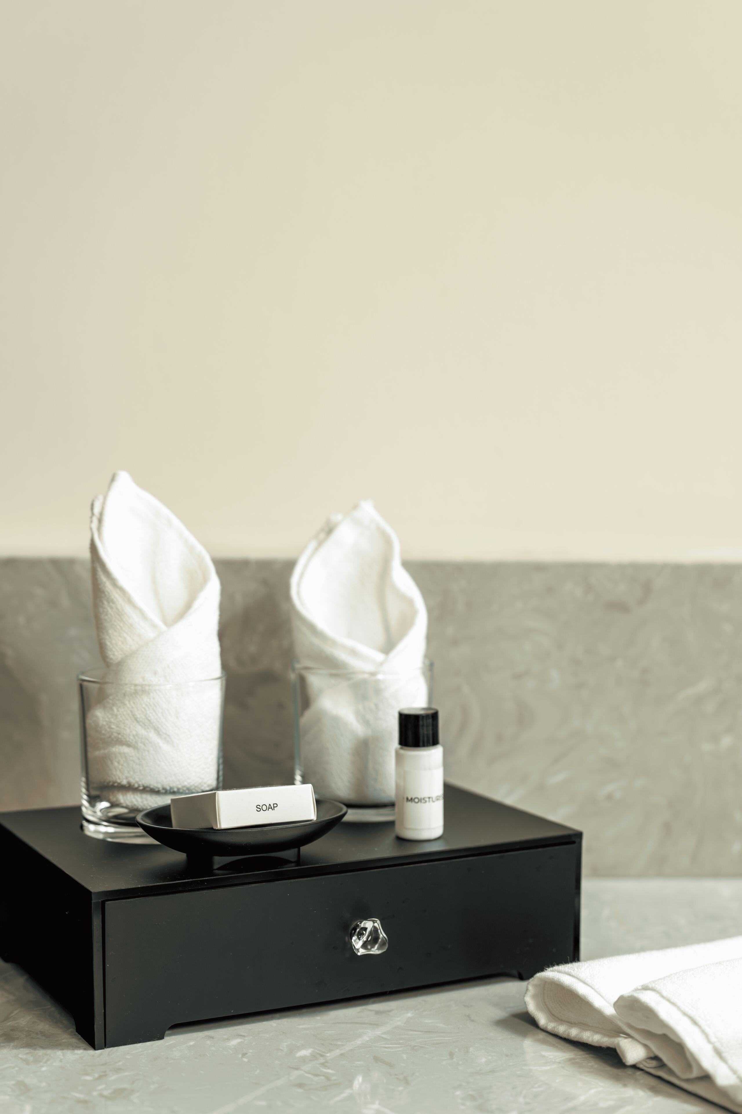 A black drawer with a soap bar, a bottle of moisturizer, and two folded towels on a marble countertop.