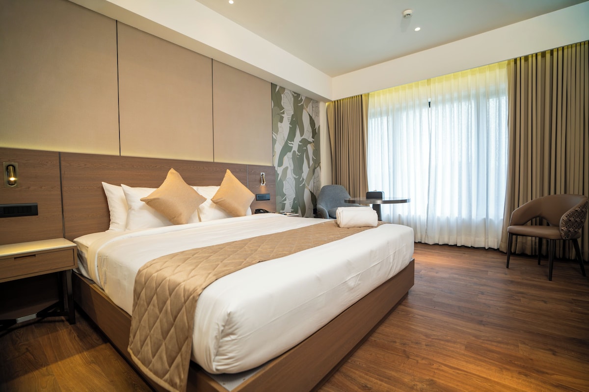 A modern hotel room with a large bed, a desk, a chair, and a view of the outdoors through sheer curtains.