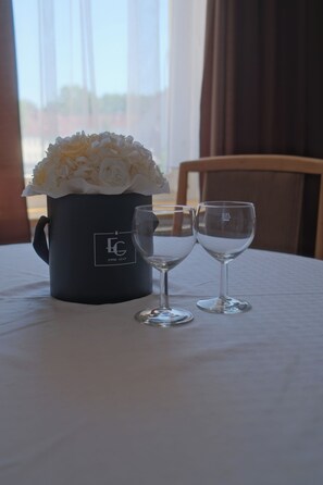 Une table avec une tasse noire et deux verres à vin, un bouquet de fleurs blanches et une fenêtre avec des rideaux.