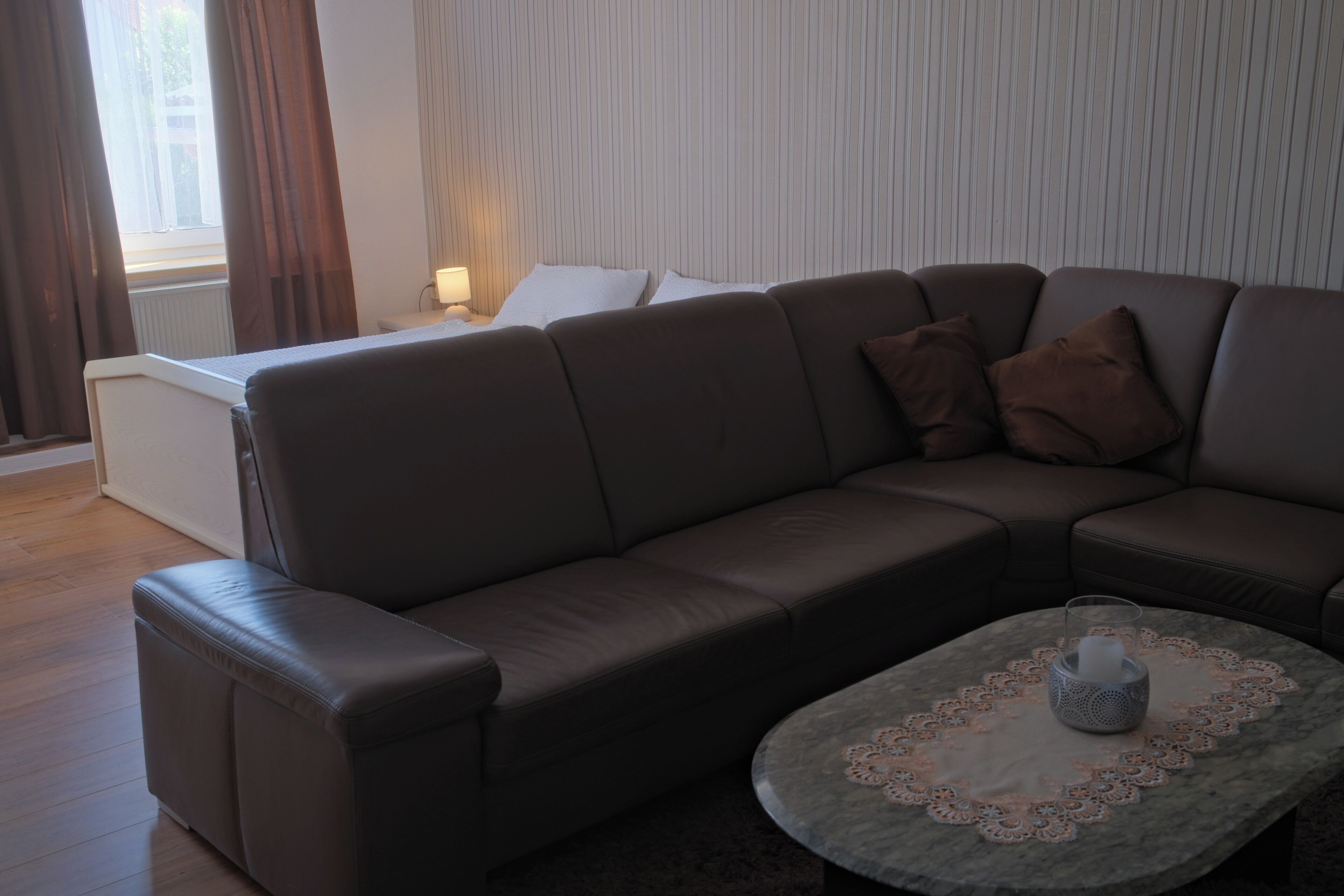 Een moderne woonkamer met een bruine leren bank, een klein rond tafeltje met een kantborduurtje, en een raam met gordijnen.