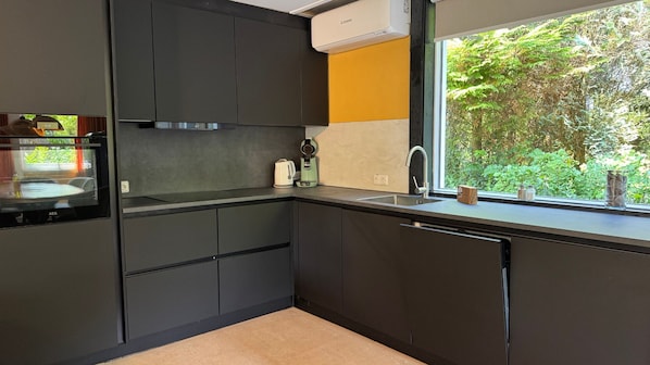 A modern kitchen with dark cabinetry, a built-in oven, a coffee maker, and a window with a view of greenery.
