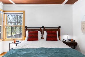 A bedroom with a bed, two pillows, a nightstand, a lamp, and a window with a view of trees. - Lundquist House | Luxury 4BR | Wraparound Deck & Hot Tub | Antler Chandelier | Idyllwild (Idyllwild)