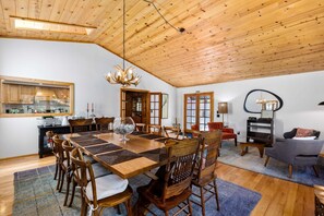Un comedor amplio con una mesa y sillas de madera, una araña y vistas a la cocina a través de una puerta de vidrio.