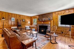 A cozy living room with a fireplace, leather sofa, and wooden walls. - Lundquist House | Luxury 4BR | Wraparound Deck & Hot Tub | Antler Chandelier | Idyllwild (Idyllwild)