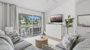 Een moderne woonkamer met een bank, een salontafel, een aan de muur bevestigde televisie en uitzicht op palmbomen door de schuifdeur.