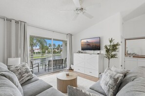 Un salon moderne avec un canapé, une table basse, un téléviseur fixé au mur et une vue sur les palmiers, visible à travers la porte-fenêtre coulissante.