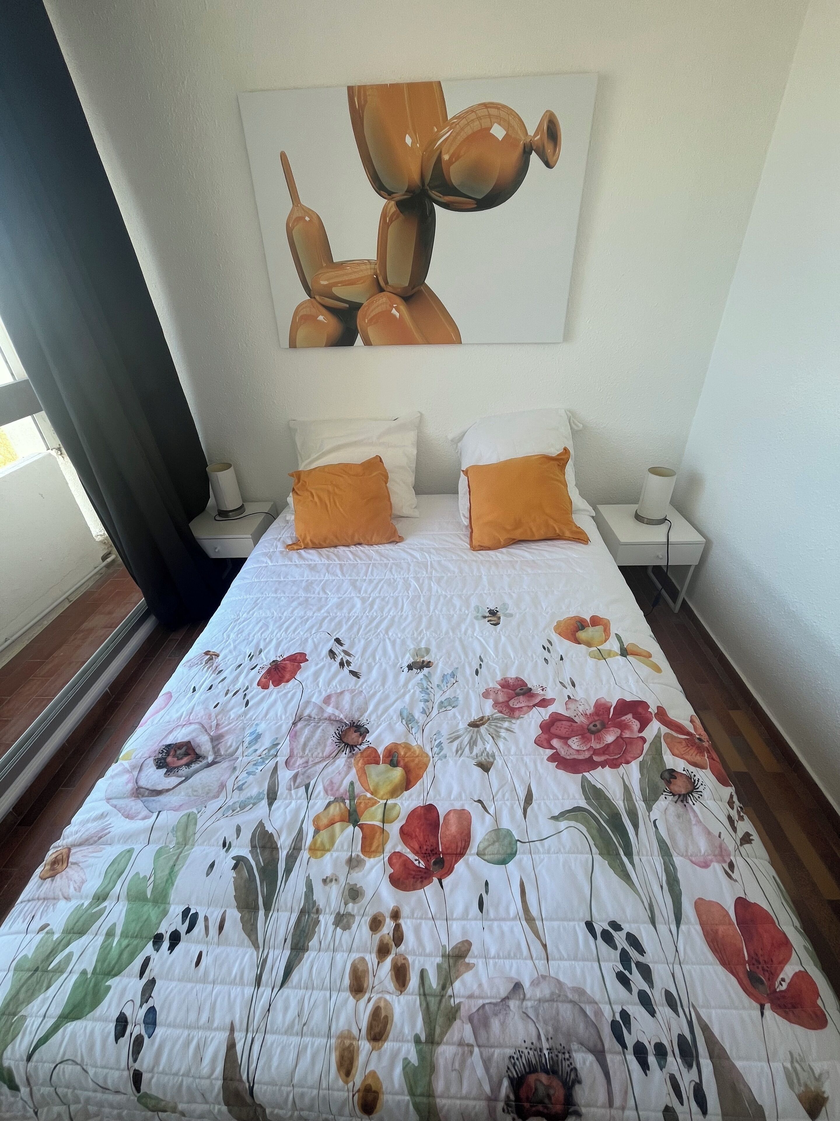 A bedroom with a floral bedspread, two white pillows, and an orange pillow. There is a painting on the wall above the bed.