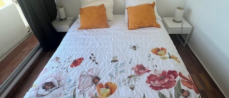 A bedroom with a floral bedspread, two white pillows, and an orange pillow. There is a painting on the wall above the bed.