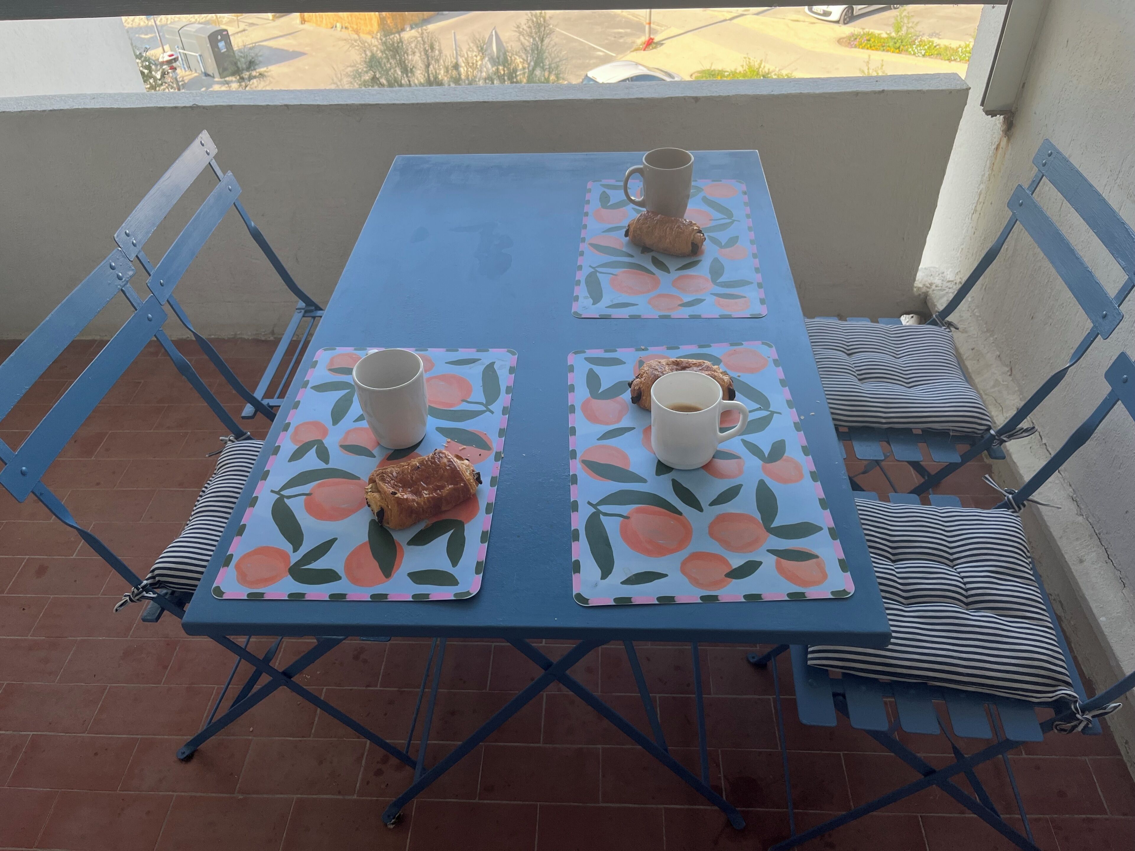 A blue metal table with two chairs, each with a cushion. The table has two placemats with a fruit pattern.