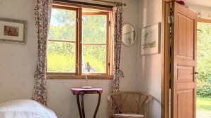 Una habitación con cama, una silla, una mesa con lámpara y un ventanal con cortinas.