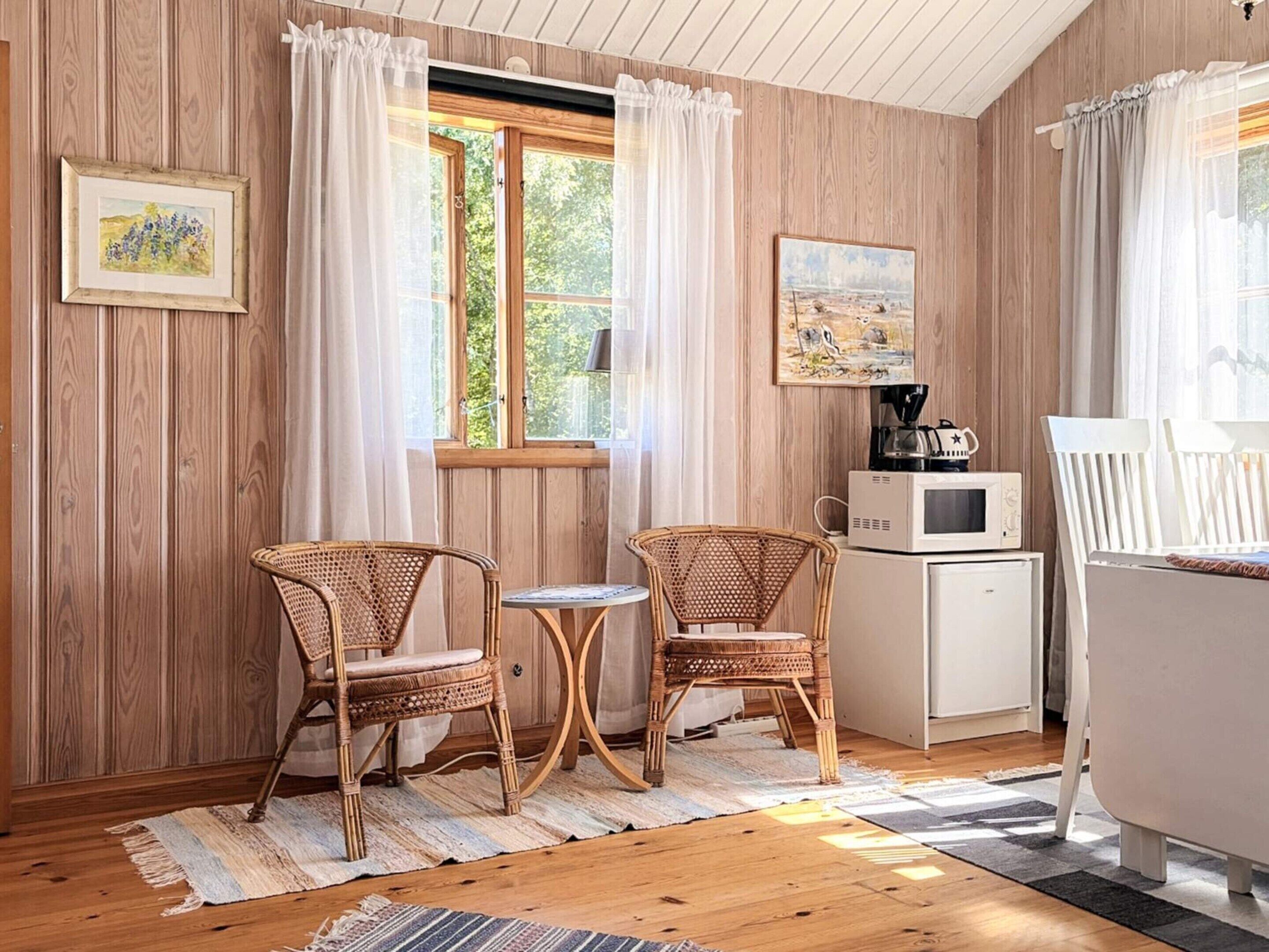 A cozy room with wooden walls, a window with curtains, two wicker chairs, a small table, a microwave, and a painting on the wall.