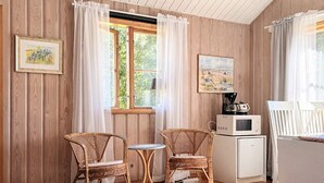 Una habitación acogedora con paredes de madera, una ventana con cortinas, dos sillas de mimbre, una mesita pequeña, un microondas y un cuadro en la pared.