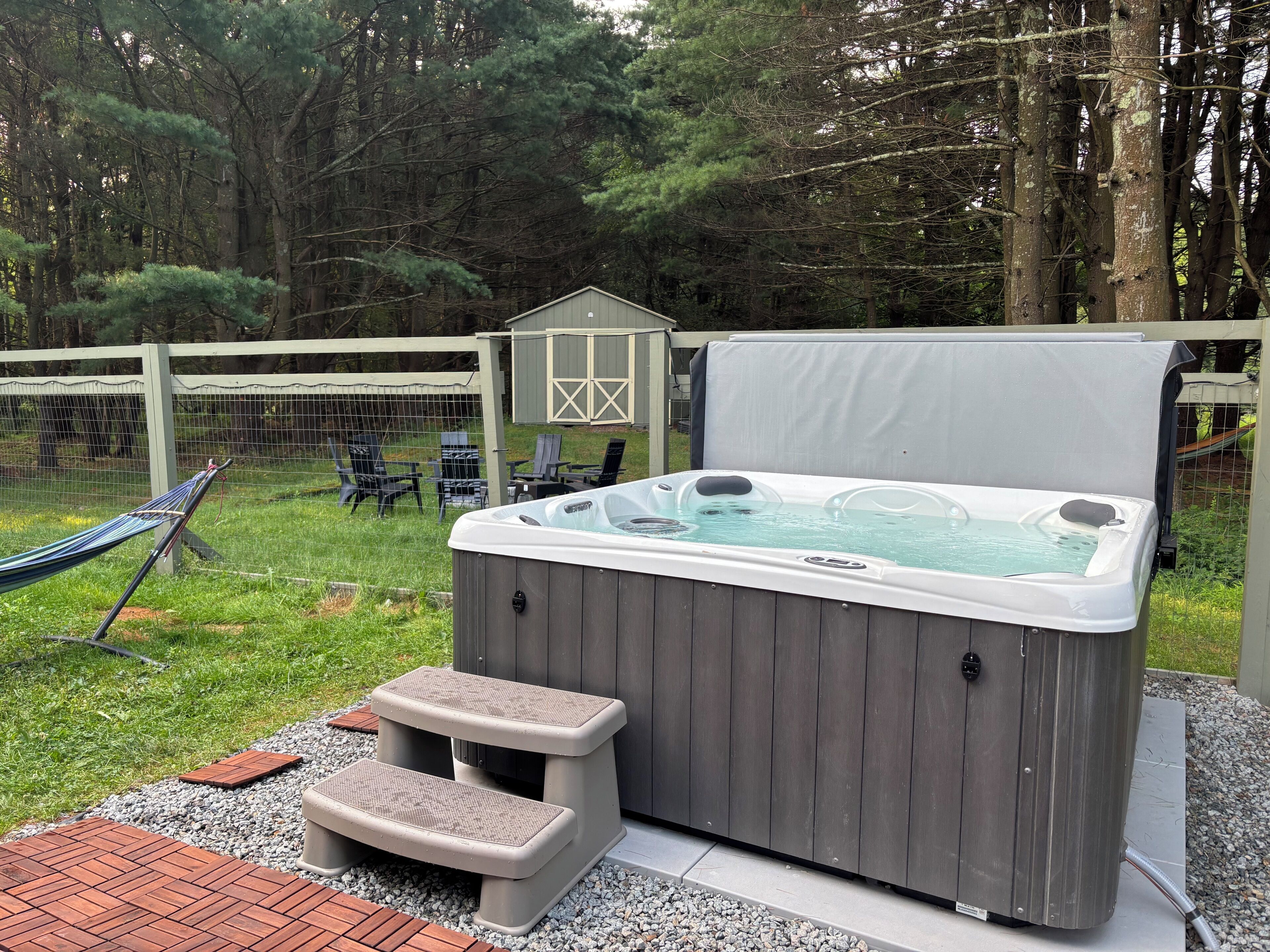A hot tub with steps, surrounded by a wooden deck, a hammock, and a fenced yard with trees.
