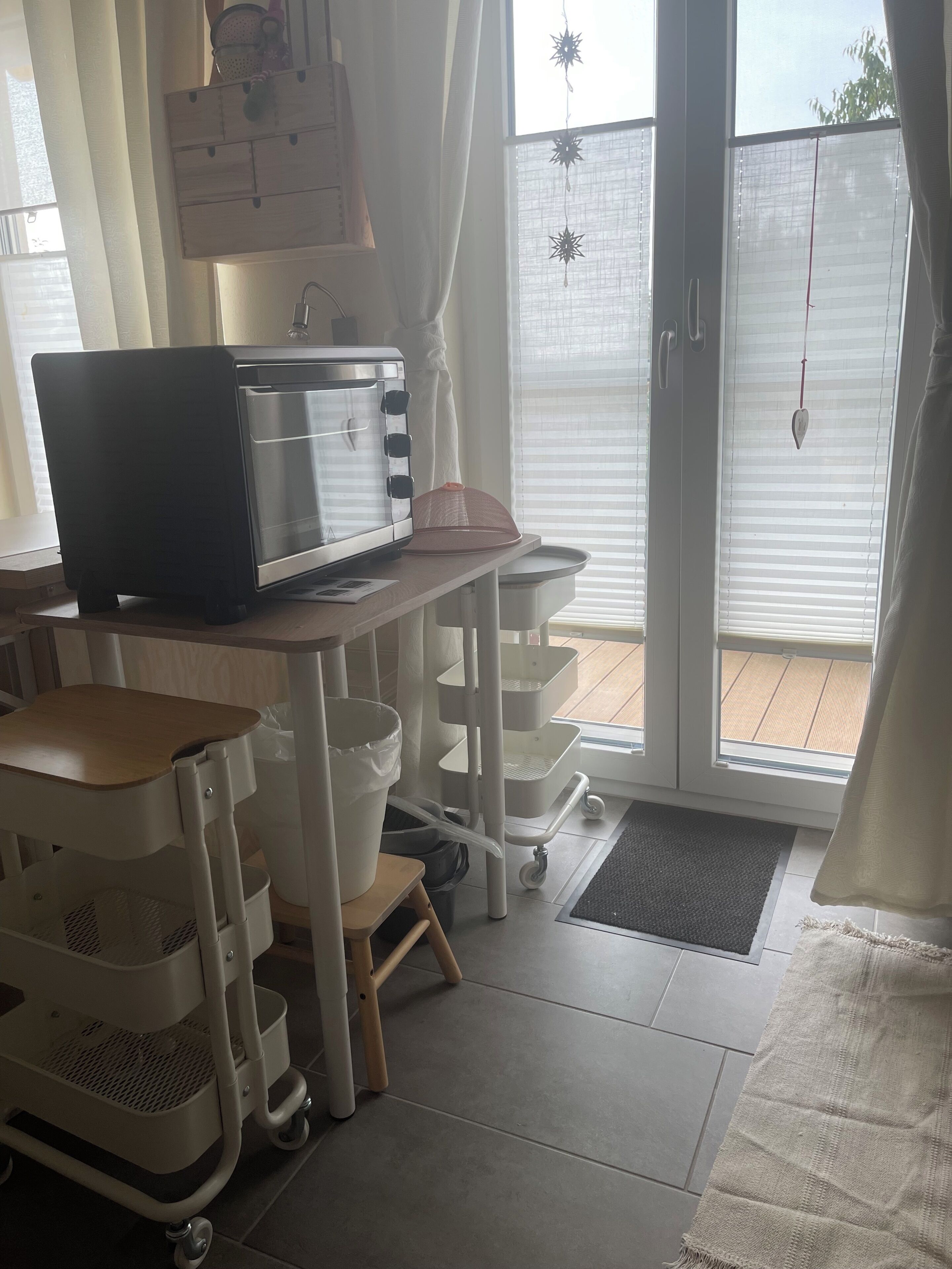 A kitchen with a microwave, a toaster, and a coffee maker on a counter. There is a window with blinds and a view of a wooden deck outside.