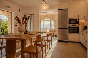 A modern kitchen with a central island, wooden chairs, and a view through an arched window. - Luxury Villa with unlimited sea view (Chania)