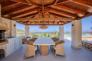 Eine Terrasse im Freien mit einem Esstisch, umringt von Korbstühlen, einem Steinkamin und Blick auf das Meer.
