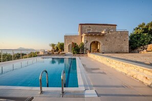 A swimming pool with a stone building in the background, a clear sky, and a view of the sea. - Luxury Villa with unlimited sea view (Chania)