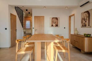 A dining area with a long wooden table and chairs, a staircase, and a kitchen area with cabinets and a fruit bowl. - Luxury Villa with unlimited sea view (Chania)