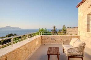 A balcony with a stone wall, a wooden table, and wicker furniture overlooking a body of water and distant mountains. - Luxury Villa with unlimited sea view (Chania)