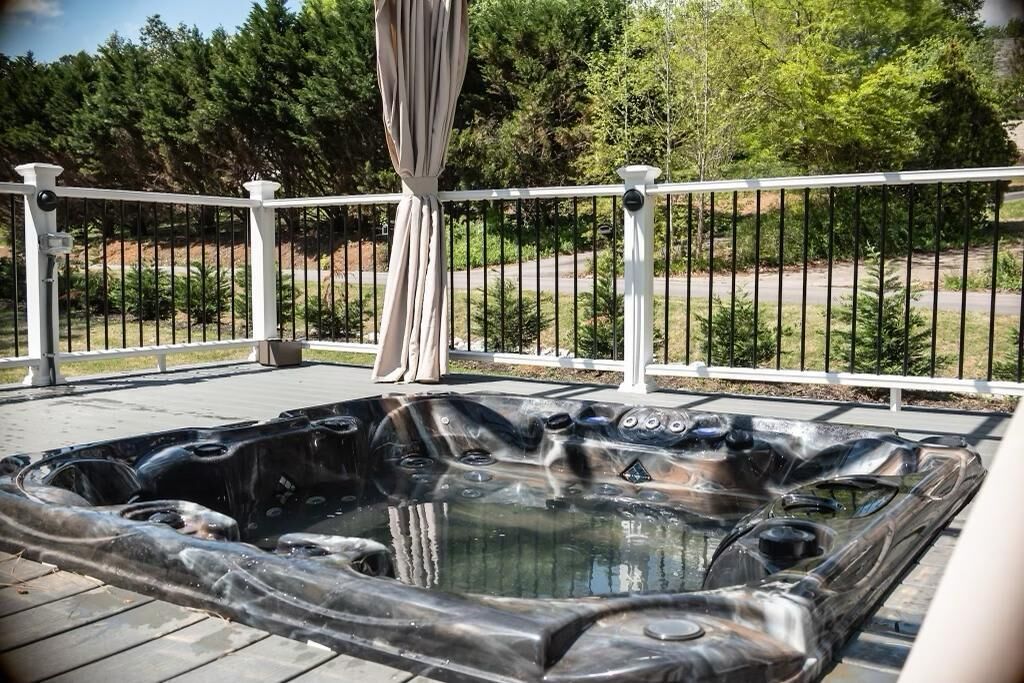 A hot tub on a wooden deck with a white railing, surrounded by greenery and trees.