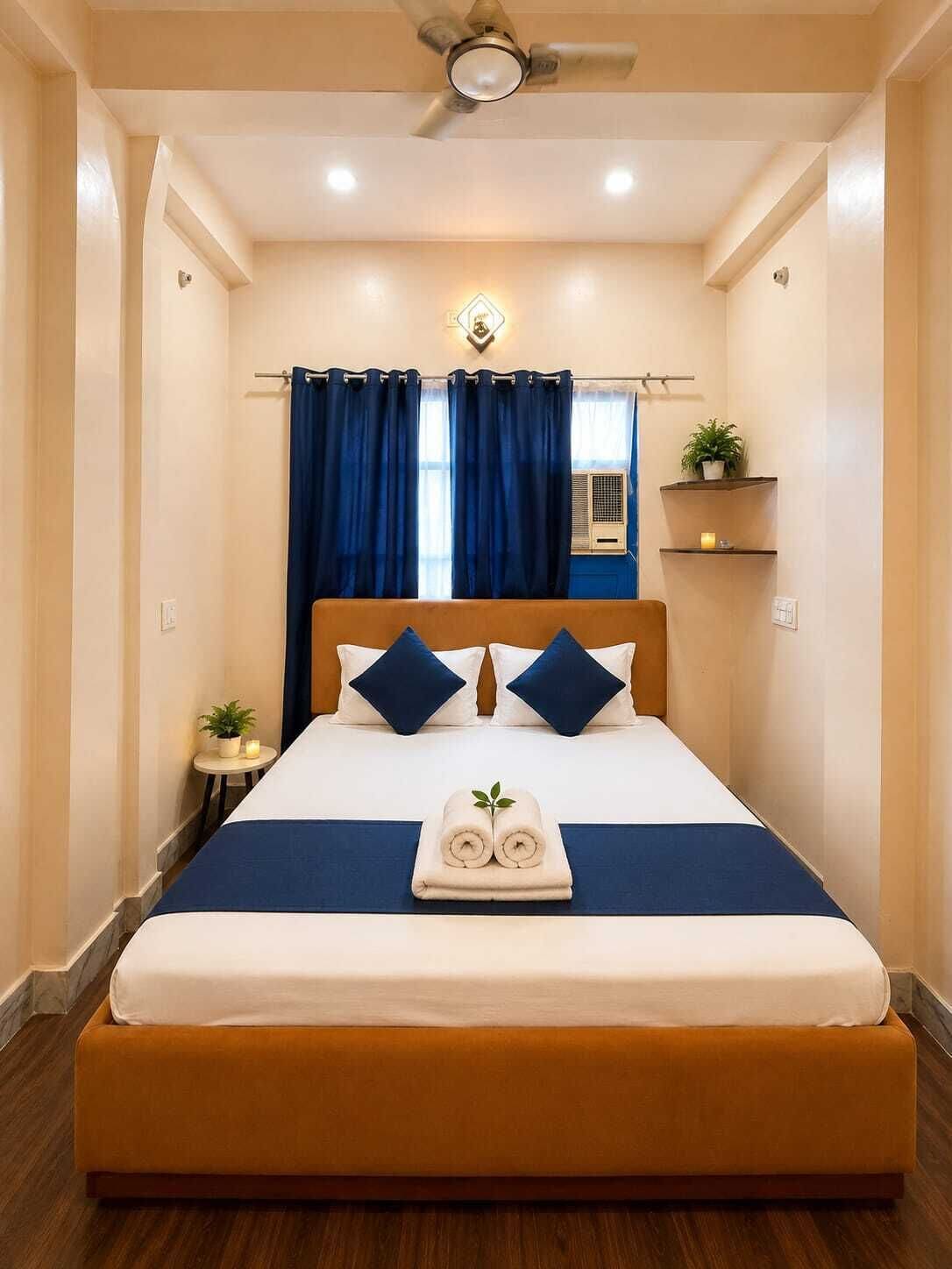 Quarto de hotel com cama, cortinas, ventoinha no teto, uma pequena mesa com uma planta e uma prateleira na parede com uma planta e uma vela.