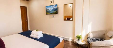 A hotel room with a bed, a chair, a small table with a plant, a painting on the wall, and a mirror.
