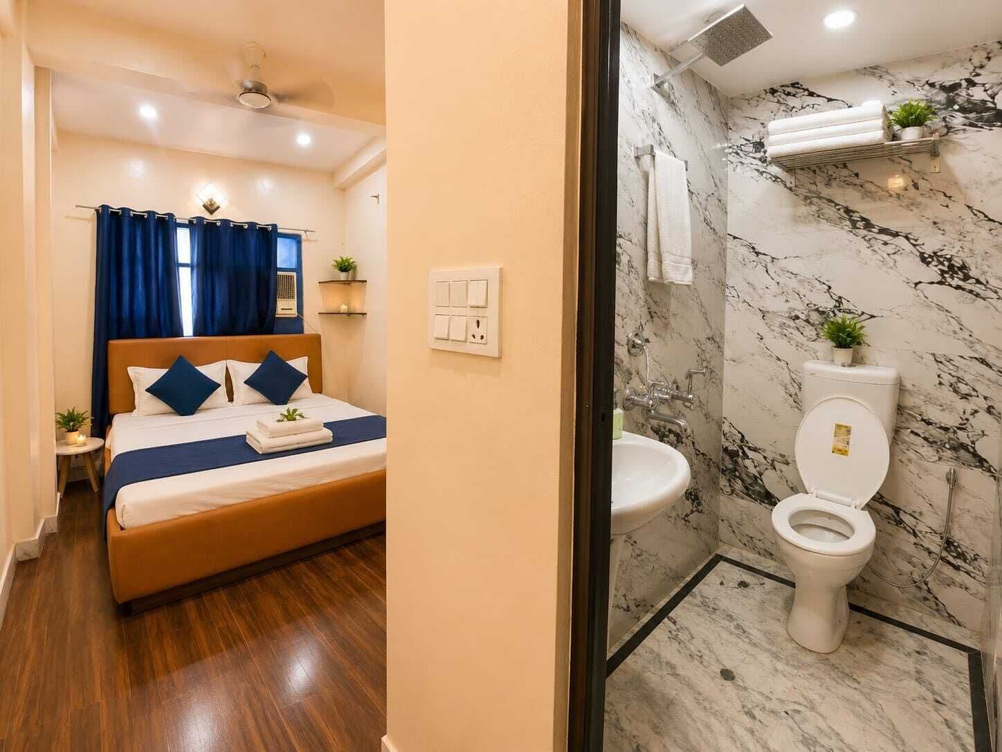 Habitación de hotel con cama, mesita de noche, ventana con cortinas, ventilador de techo y baño con inodoro, lavabo y paredes de mármol.